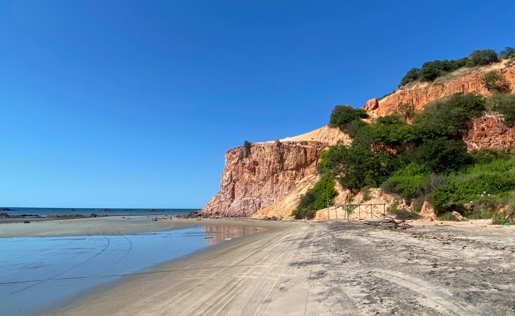 An orange rocky formation surrounded by vegetation at the beach with the sea to its left. The sky is clear and deeply blue.