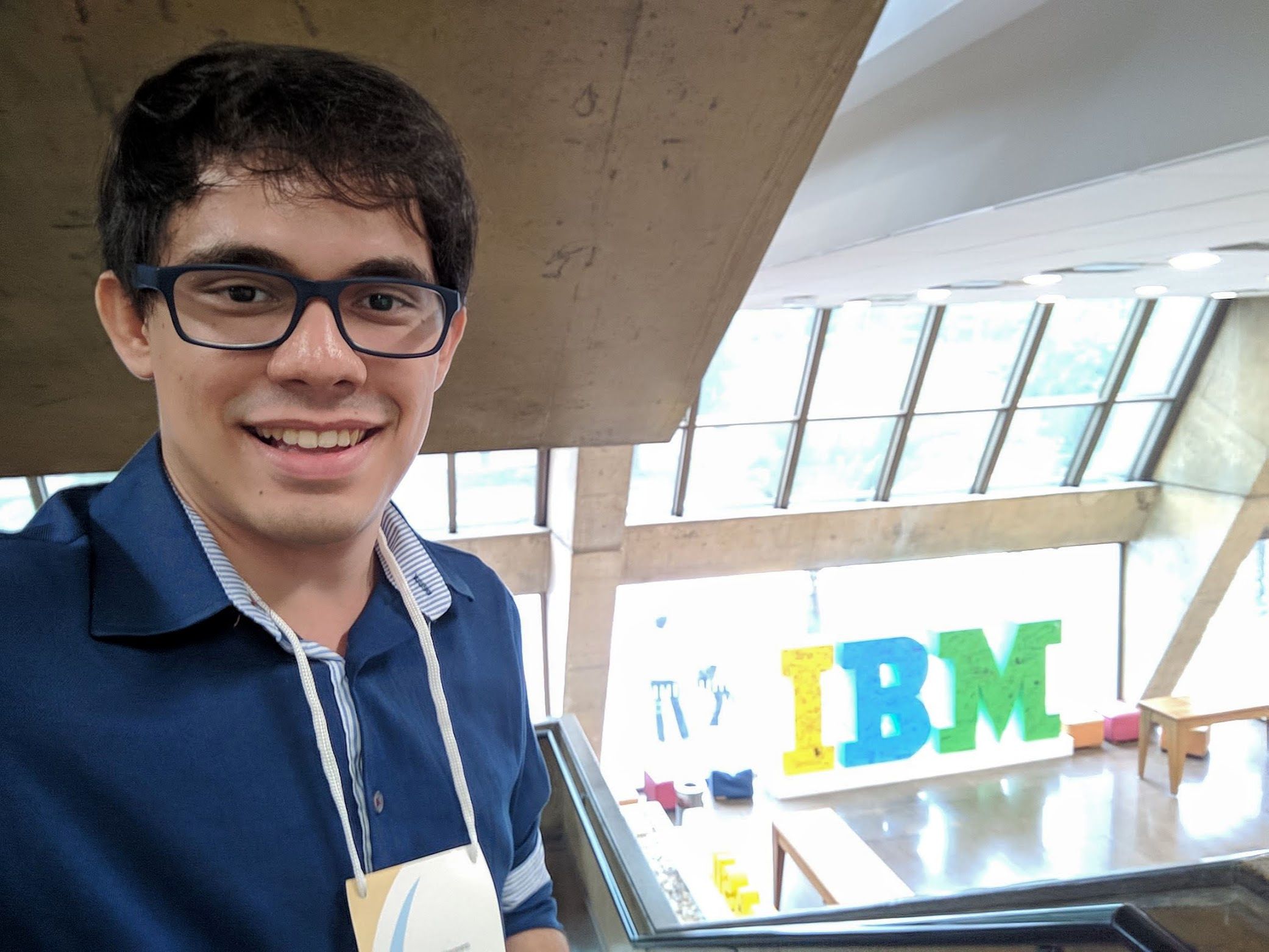 A selfie taken by me with the IBM Brazil entry hall as background. I wear squared glasses and a blue dressing shirt, with a conference badge placed on my neck. My hair is straight and combed to the left.