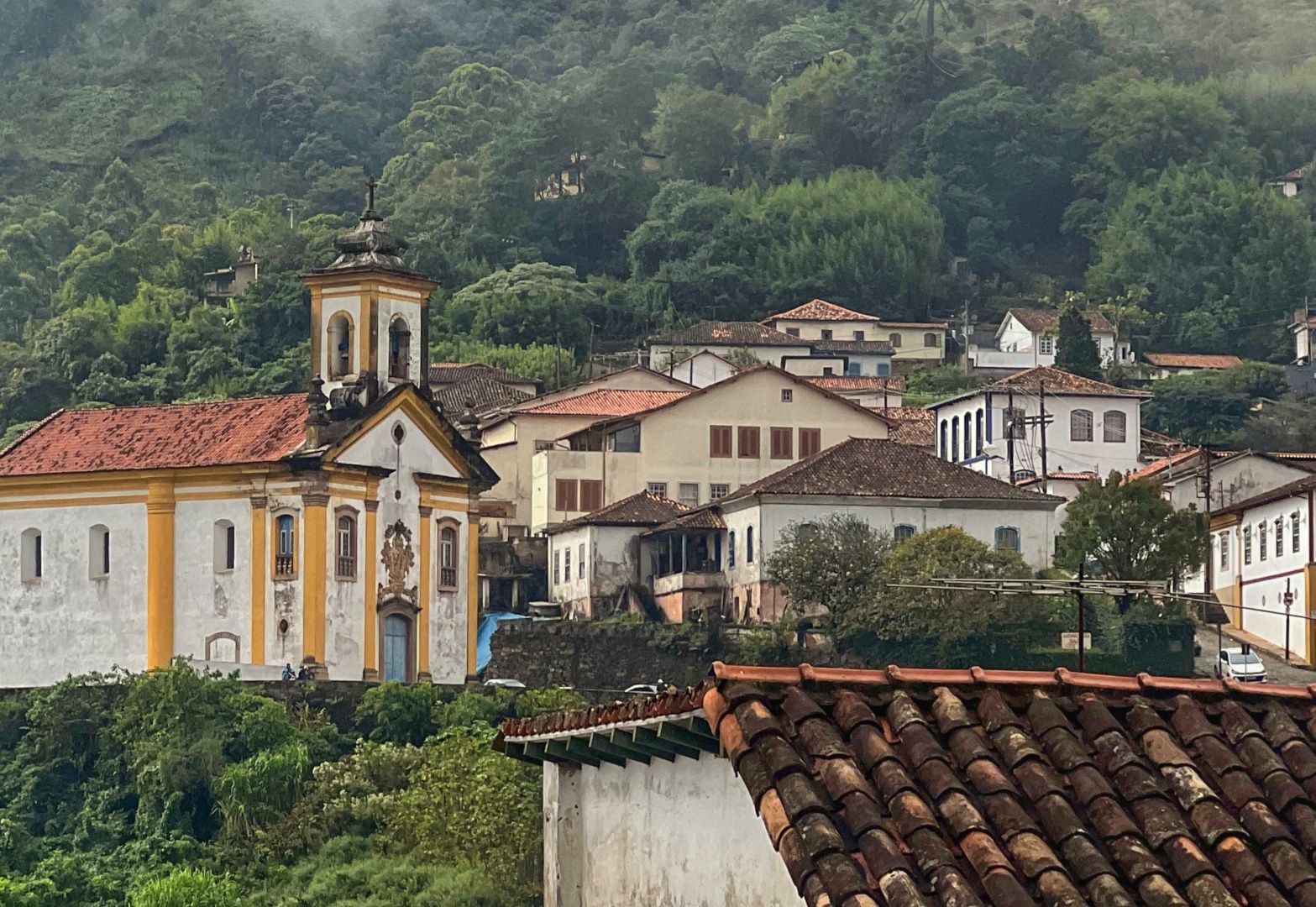 A historical church in a hill in Ouro Preto. The church, built in Baroque architecture style, has a central tower and two windows in its façade. To its right, a street surrounded by colonial style households goes up the hill.